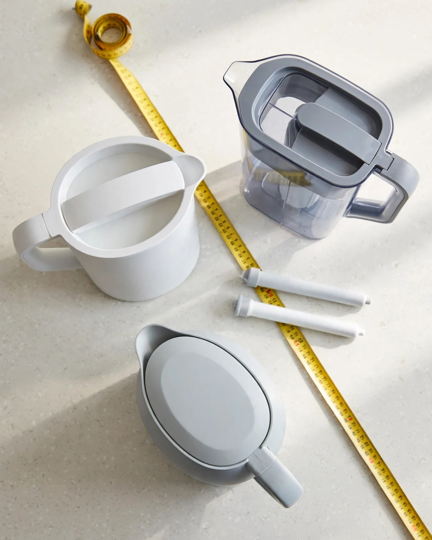 Overhead flat-lay of three water filter pitchers and two filter cartridges with a yellow measuring tape on a light kitchen counter