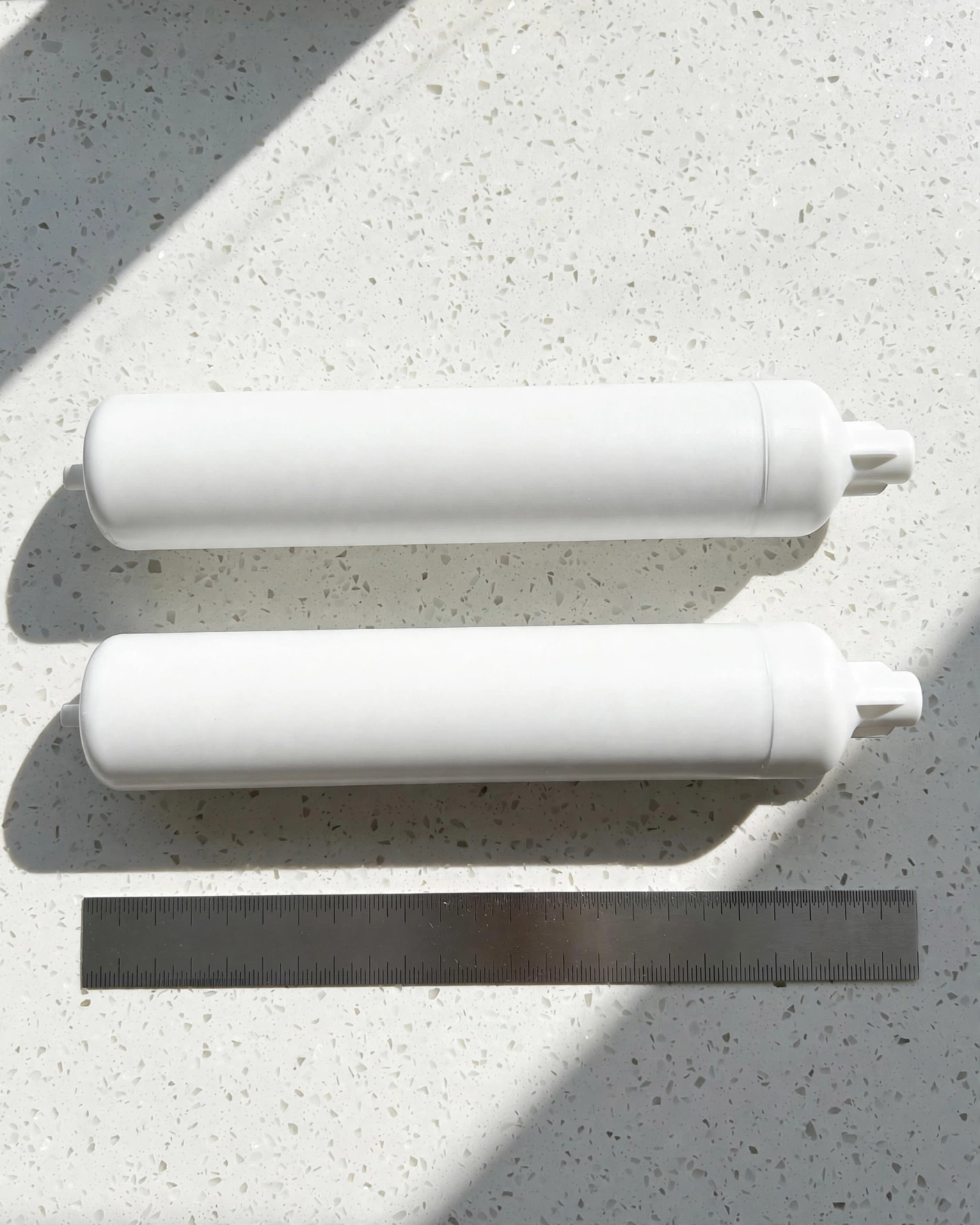 Two unlabeled fridge filter cartridges with a steel ruler on a quartz counter — checking exact fit dimensions